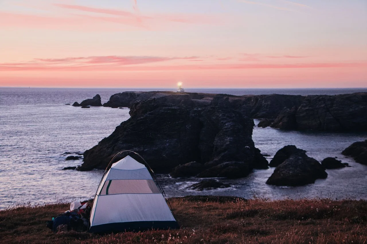 A small tent pitched on a grassy headland above a quiet Atlantic cove at sunset