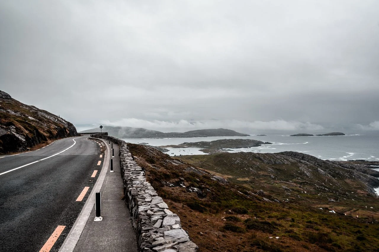 A narrow Irish coastal road winding along green Atlantic headlands under a moody sky